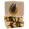 Three stacked rectangular bars of handmade soap on a plain white background. The top bar is light beige with a stylized flame-like logo in green, mustard yellow, and dark burgundy.